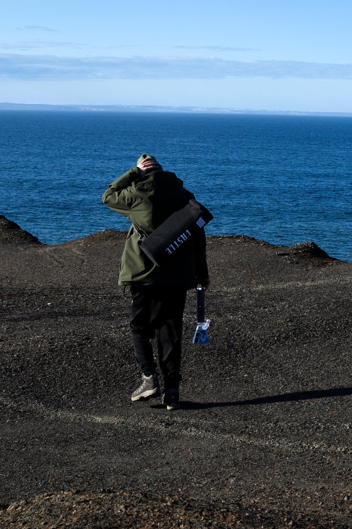 A person in a green jacket and black pants stands on a rocky, barren landscape overlooking the ocean. They have their back to the camera and are holding a clear glass bong in one hand, while a black carrying bag with the "CASTLE" logo slung over their shoulder. The bright blue sea stretches out to the horizon under a clear sky, creating a striking contrast with the dark ground.