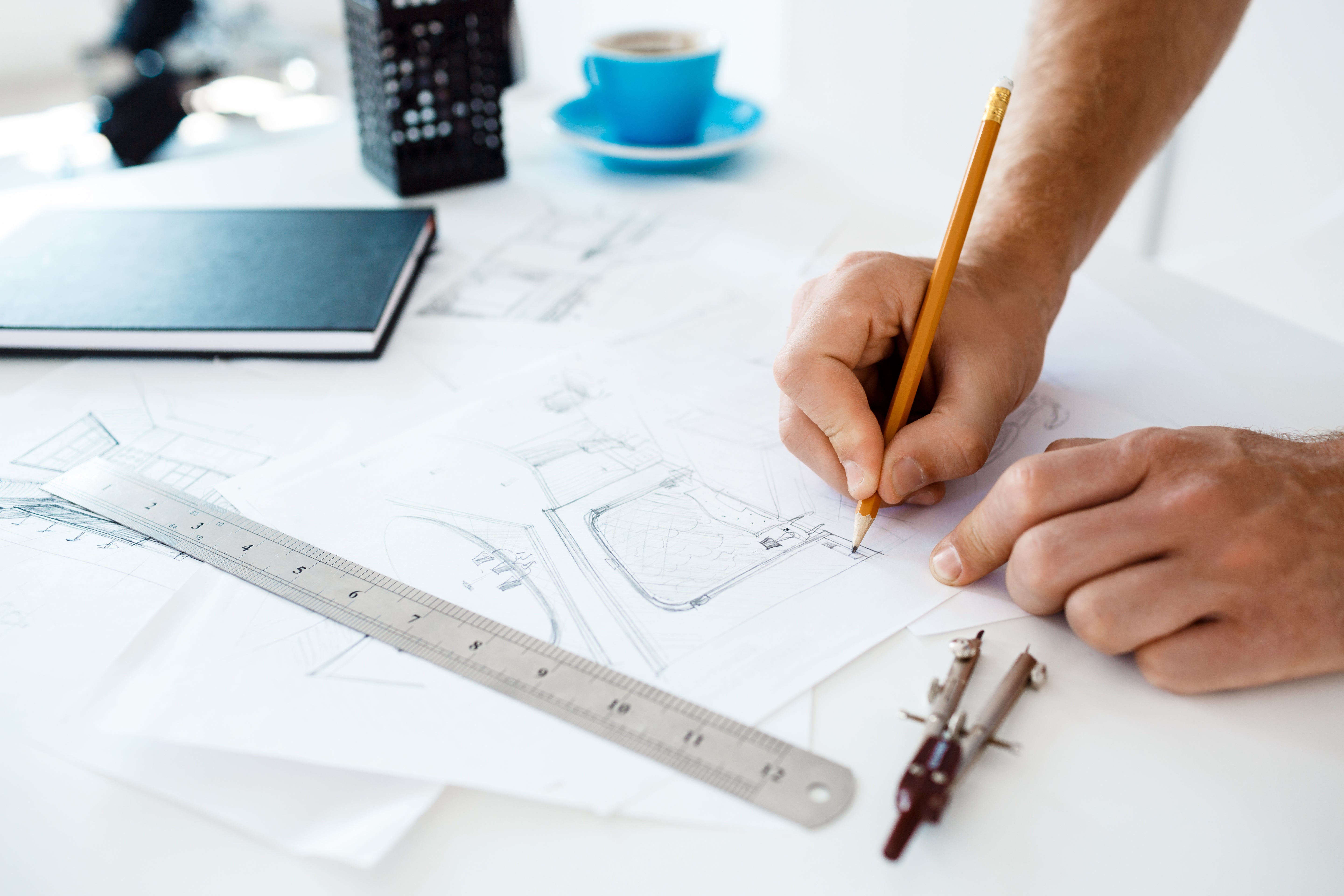 Close-up of a person’s hands drawing architectural sketches with a pencil on white paper, alongside a ruler, compass, and a notebook.